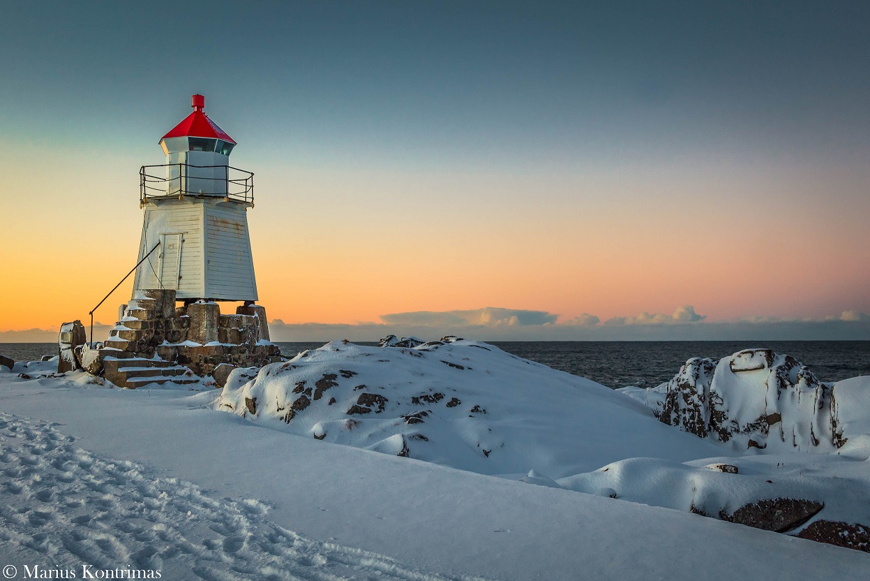 Fyrlykt på snødekt holme i Lofoten i solnedgang, med åpent hav i bakgrunnen